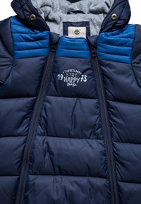 Marinblå puffjacka med huva, med kviltad textur, blå accenter och dragkedja fram. Visar "Timberland Stay Happy 73"-logotyp.