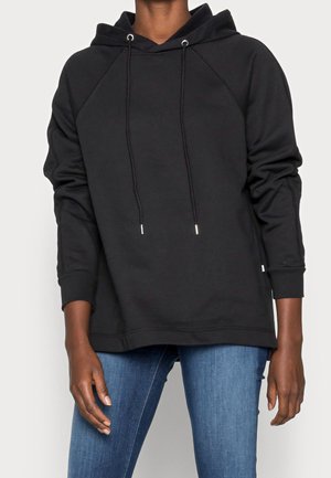 Sort langærmet hættetrøje med snore, båret med blå jeans, viser overkroppen og afslappet armstilling.