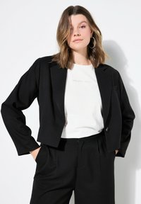 Blazer nero corto con maniche lunghe, chiusura a un bottone e tasca frontale. Indossato sopra una camicia bianca con testo, abbinato a pantaloni neri.