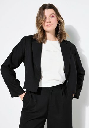 Schwarze, taillierte Blazerjacke mit langen Ärmeln, einreihigem Knopfverschluss und Fronttasche. Getragen über einem weißen Hemd mit Aufdruck, kombiniert mit schwarzen Hosen.