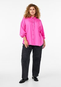 Ružičasta oversized košulja s dugim rukavima, ovratnikom, jednim džepom na prsima i zavrnutim rukavima, uparen s crnim ravnim trapericama i crnim cipelama.