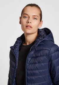 Marineblauwe gewatteerde jas met capuchon, gestikt ontwerp en ritssluiting. Draag het over een zwart T-shirt; de stof lijkt glanzend met een gladde textuur.