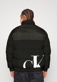 Sort dunjakke med vandrette paneler. Har en høj krave og en hvid logo på ryggen. Glat tekstur, ribstrikkede manchetter og kant.