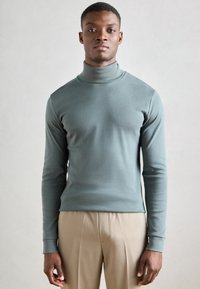 Gai zila garām piedurknēm turtlenecka stila džemperis, izgatavots no mīksta materiāla, ar pieguļošu piegriezumu un ribotiem piedurkņu galiem. Valkāts kopā ar bēšām biksēm.
