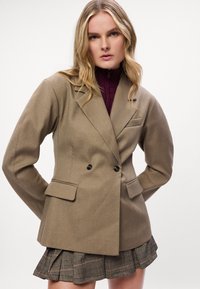 Blazer castanho de dupla face em tecido texturizado com lapelas recortadas, bolsos frontais e botões pretos, usado sobre uma saia plissada xadrez.