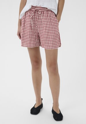 Ben iført røde og hvide ternede shorts med snøre i taljen, kombineret med sorte slip-on sko og en hvid top.