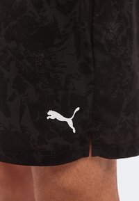 Μαύρα σορτς με σχέδια και λευκό λογότυπο Puma κοντά στο τελείωμα, φορεμένα από άτομο με ορατή τριχοφυΐα στα πόδια.