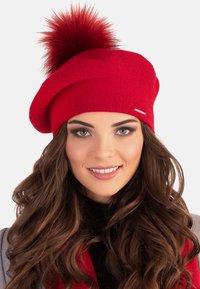 Berretto rosso lavorato a maglia con un grande pompon in pelliccia sintetica sulla parte superiore, caratterizzato da una superficie liscia e un piccolo logo metallico decorativo.