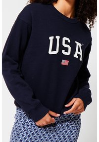 Marinblå sweatshirt med "USA" i vit skrift och en liten amerikansk flagga-patch. Har långa ärmar och rundad halsringning, tillverkad av mjukt material.