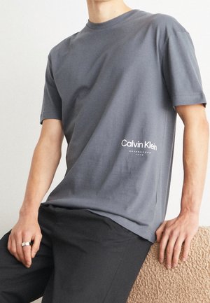 Person iført en grå Calvin Klein T-shirt med logo i nederste højre side, sorte bukser og en sølvring på venstre hånd, siddende på en tekstureret overflade.