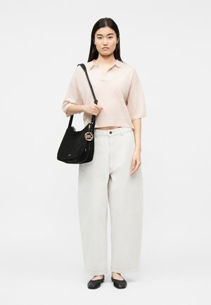 Žena u svijetloružičastoj majici kratkih rukava, bijelim širokim hlačama, crnim baletankama i s crnom torbom na rame sa zlatnim privjeskom.