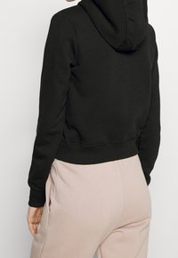 Schwarze cropped Kapuzenjacke mit einer Fronttasche, gerippten Bündchen und Texturdetails. Getragen mit hellrosa Jogginghosen, die ein lässiges Outfit präsentieren.