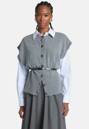 Femme portant un gilet en maille gris boutonné avec une ceinture sur une chemise blanche à manches longues et une jupe grise, debout devant un fond uni.