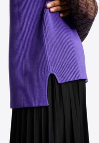 Lila gerippter Pullover mit asymmetrischem Saum und seitlichem Schlitz. Die Textur ist weich und gestrickt, kombiniert mit einem plissierten schwarzen Rock.