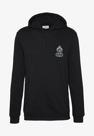 Zwarte hoodie met groene grafiek, zwarte heuptas, grijze spijkerbroek, en rode hoge sneakers met witte zolen.