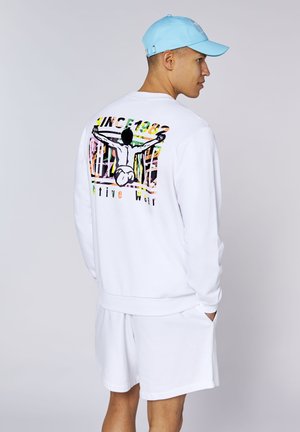 Mann trägt weißen Pullover und Shorts, mit Grafik einer Person, die die Arme auf der Rückseite des Pullovers ausstreckt, hellblaue Kappe, Hände in den Taschen.