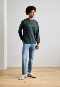 Sweatshirt vert foncé avec un col rond, arborant un texte logo blanc. Associé avec un jean bleu clair et des sneakers verts, blancs et marron.