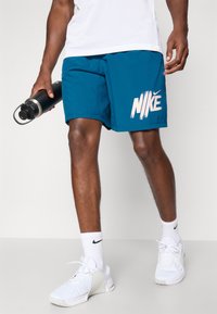 Türaun Nike-Shorts in Teal mit weißem Logo, aus feuchtigkeitsableitendem Stoff, mit elastischem Bund, kombiniert mit weißen Nike-Schuhen und einer schwarzen Wasserflasche.