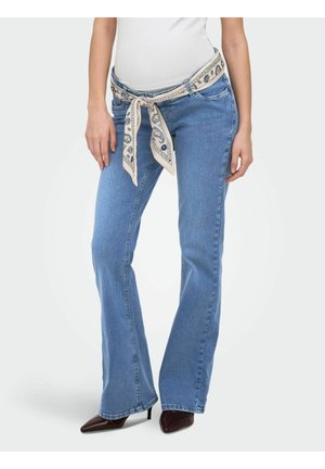 Vrouw draagt lichtblauwe bootcut jeans, witte crop top, bruine puntige hakken en een beige sjaal met patroon, gestrikt als riem.