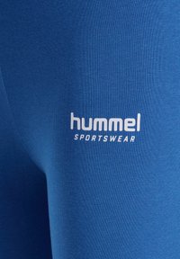 Niebieskie legginsy sportowe wykonane z elastycznego materiału, z białym logo "hummel" i napisem "SPORTSWEAR" poniżej, w pobliżu dołu.