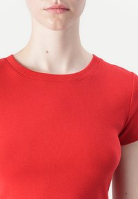 Punane ja tihe lühikeste varrukatega topp ümmarguse kaelusega. Kude on sile ja ribakujuline, näidates standardset siluetti ilma mustriteta.