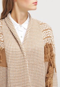 Beżowy ręcznie szyty szal z białymi wzorami, z frędzlami na krawędziach i fakturowanym wykończeniem. Noszony na białej koszuli z kołnierzykiem.