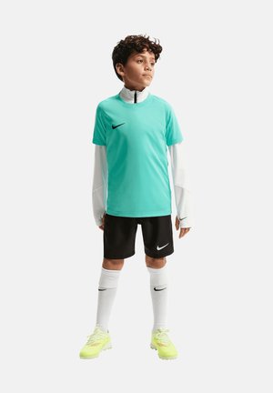Niño con camiseta deportiva turquesa sobre camiseta blanca de manga larga, pantalones cortos negros, calcetas blancas hasta la rodilla y botas amarillas, de pie mirando a la derecha.