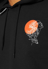 Sudadera negra con un dibujo lineal en blanco de un rostro con cabello fluido, superpuesto por una forma circular naranja. Se aprecia una textura de tela suave.