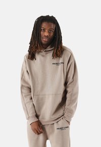 Sudadera con capucha beige, con un bolsillo frontal y el logo negro "PROJECT X PARIS". Textura suave y ajuste holgado, combinada con pantalones a juego.