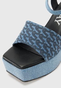 Denimplattformssandal med ett blått mönstrat band, tår i svart läder, och en texturerad bas i blå denim. Fyrkantig form med en öppen tå-design.