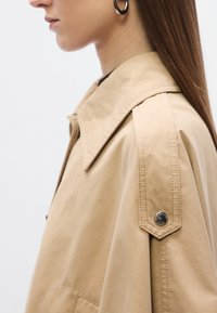 Bež, velika košulja jakna s velikim ovratnikom, naramenicama i srebrnim kopčama na drukere. Glatka tekstura tkanine.