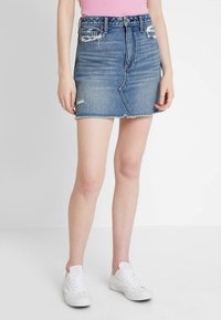 Jeans-Minirock, mittlere Blautonwäsche, abgenutzte Details, ausgefranster Saum, hoch tailliertes Design, kombiniert mit weißen Turnschuhen.