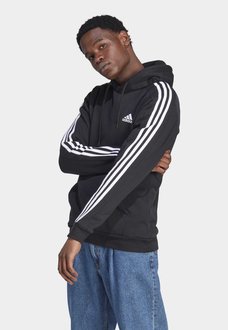Sudadera con capucha negra y diseño de tres rayas blancas a lo largo de las mangas, con un logo de Adidas en el pecho. Material suave, corte relajado.