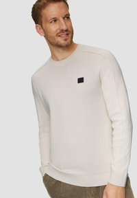 Jasno-beżowy sweter dzianinowy z okrągłym dekoltem i ściągaczami. Posiada małą czarną naszywkę z logo na lewej piersi. Teksturowany materiał.