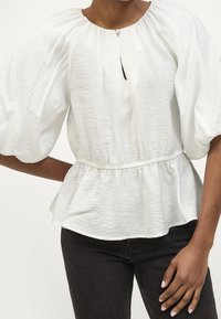 Blouse blanche légère avec des manches bouffantes, une taille froncée et un détail en forme de clé au niveau du col. Texture lisse avec une coupe fluide.