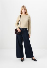 Lys blond kvinne iført beige genser, vide mørke jeans, svarte leiligheter og en liten mørkebrun crossover-veske som står mot hvit bakgrunn.