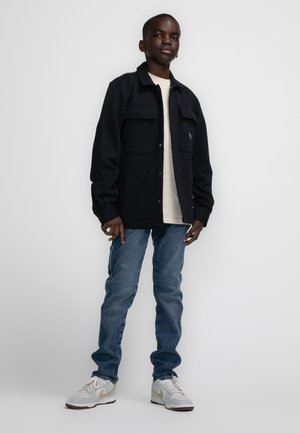 Zwarte buitenoverhemd met twee borstzakken, gedragen over een lichtgekleurde shirt. Blauwe jeans en grijze sneakers met gouden accenten maken de outfit compleet.