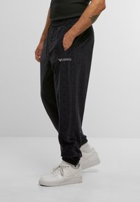 Man wearing black textured Colucci sweatpants, white socks, and white Nike sneakers, standing with hands in pockets against a plain background.