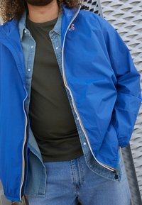 Blauw lichtgewicht jack met rits en meerkleurige bies, gedragen over een groen shirt en lichte spijkerbroek, met zichtbare knoopdetails.