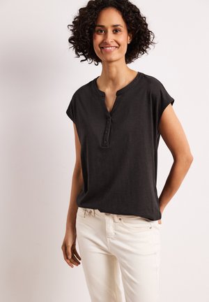 Mujer con el pelo rizado que lleva una camiseta Henley negra de manga corta y pantalones blancos, sonriendo con una mano en el bolsillo.