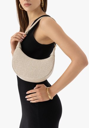 Frau in schwarzem Kleid, die eine beigefarbene gewebte Umhängetasche hält, mit goldenen Creolen und goldenen Armbändern vor weißem Hintergrund.