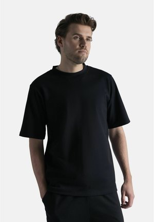 Mies, jolla on lyhyet ruskeat hiukset, pukeutuneena yksinkertaiseen mustaan lyhythihaiseen t-paitaan, seisoo valkoisen taustan edessä, kädet osittain taskuissa.