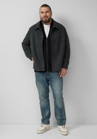 Donkergrijze button-up jas over een zwarte ribbeltrui, gecombineerd met lichtblauwe jeans en witte sneakers. Glad texturen en effen kleuren.
