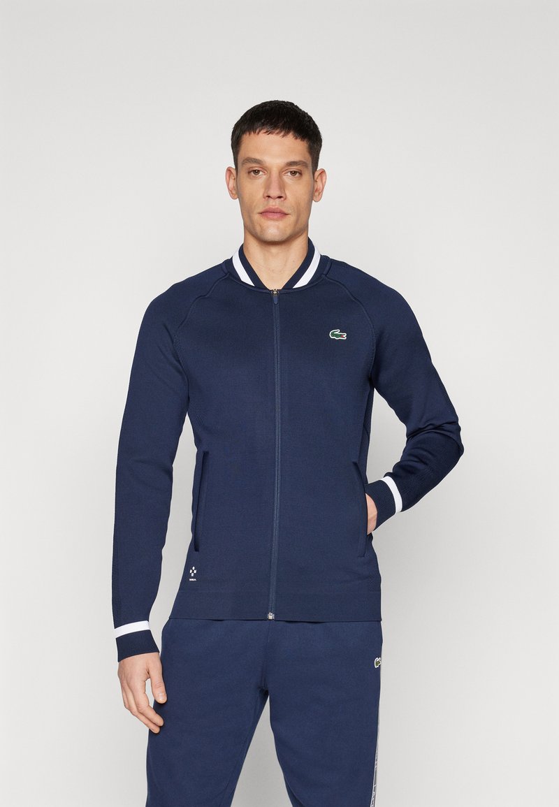 Casaco azul marinho com fecho éclair e gola com riscas brancas, equipado com um emblema, punhos canelados e bolsos laterais. Textura suave e design ajustado.