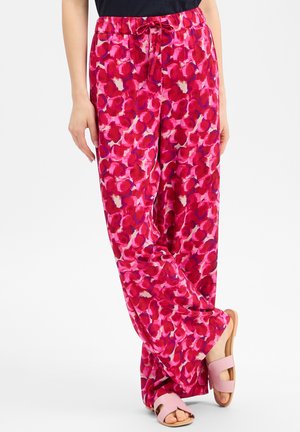 Pantalon ample avec un motif floral rouge et rose, taille élastique avec cordon, porté avec des sandales ouvertes roses.
