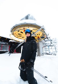 Skiing equipment and black outerwear worn by a person standing in snow near a ski lift, featuring yellow gears and metal chairs in background.
