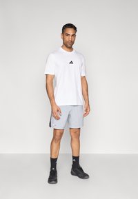 Camiseta deportiva blanca con logo negro, combinada con pantalones cortos gris claro con franjas laterales negras. Medias negras y zapatillas completan el conjunto.
