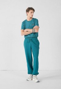 Teal katoenen t-shirt met een klein logo, gecombineerd met bijpassende joggingsbroek met een elastische tailleband en koord, en grijze sneakers.