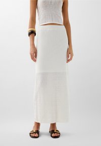 Witte, gebreide maxi rok met textuurpatronen; aansluitende taille, lichte stof; gedragen met donkere slidesandalen met een gouden accent.