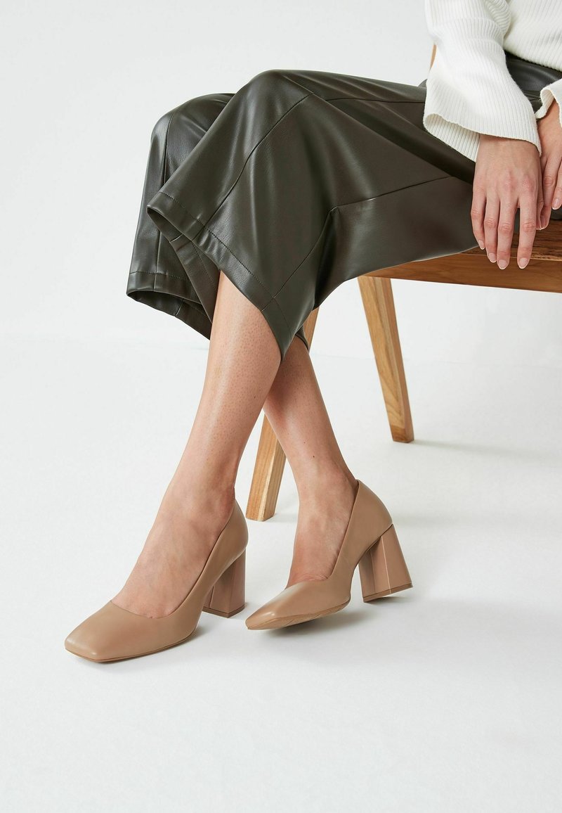 Escarpins à talons hauts en cuir verni beige, avec bout carré et talon épais, portés avec une jupe en cuir foncé et posés sur un fond minimaliste.